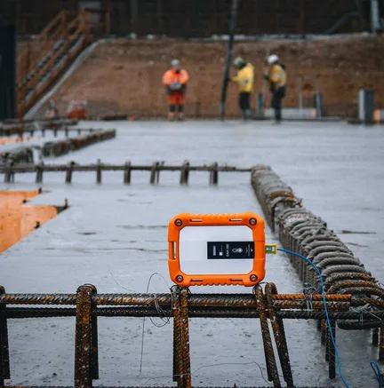 Orangefarbener Sensor mit Kabel auf einer frisch betonierten Fläche, im Hintergrund Bauarbeiter