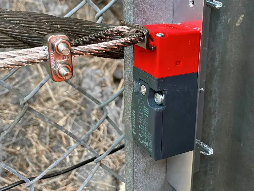 Seilzugsensor mit rotem Gehäuse am Zaunpfosten, verbunden mit dem Drahtseil der Auffangbarriere